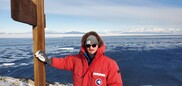 A man in a big red jacket smiles at the camera while standing on ice with the ocean in the background.