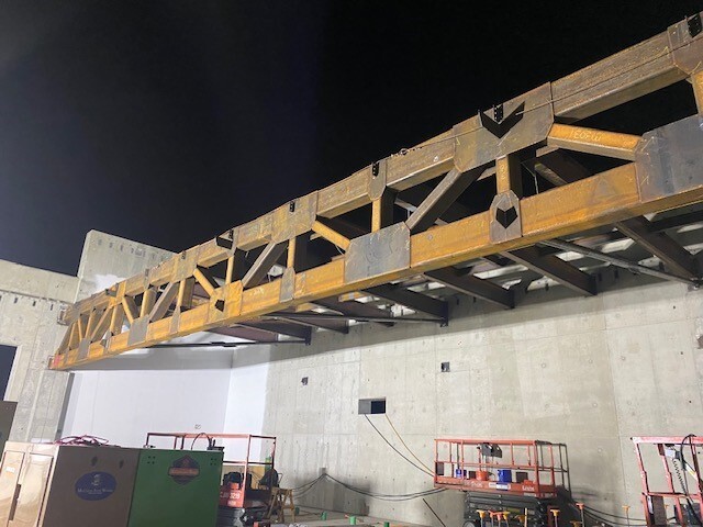 An 86 foot long truss is lowered into place to start forming the roof of a concrete building. 