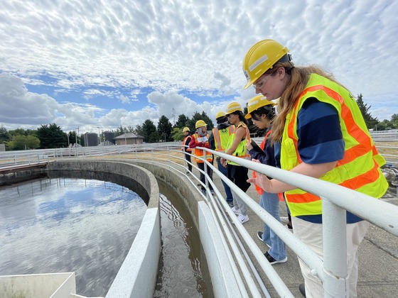 A group of young people in hardhats, safety glasses and vests look over a rail at a wastewater treatment plant. 