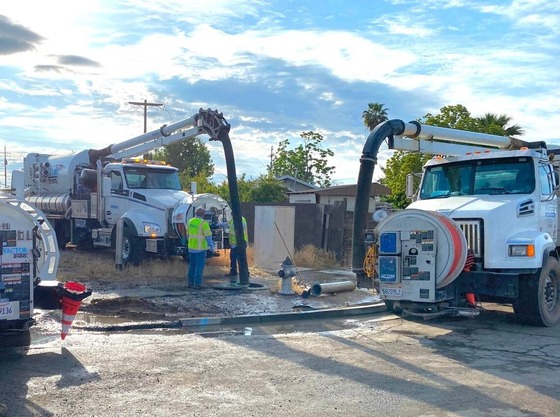 Two workers wearing neon safety shirts stand in the middle of three vactor, or vacuum, trucks.  
