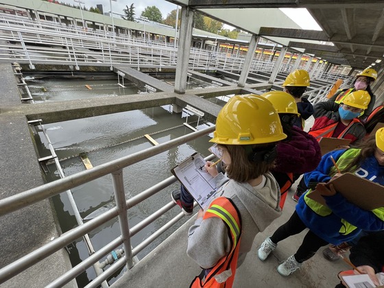 School children wearing hard hats, safety glasses and neon safety vests write on clipboards at a wastewater treatment plant. 
