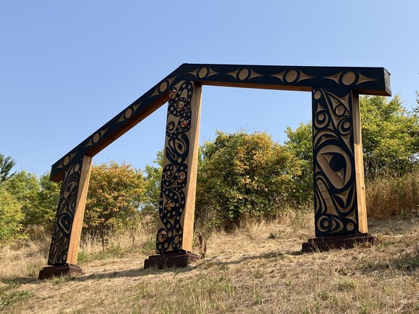 An old-growth cedar sculpture featuring carvings that honor Coast Salish peoples and traditions pn the side of a grassy hill.