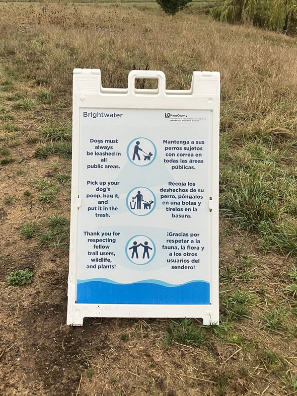 A white A frame sign stands next to a trail. The sign reminds trail users to leash their dogs and pick up and dispose of their dog's poop.