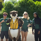a photo of a person wearing an otter mascot costume with three other teens smiling at the camera
