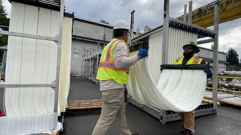 Two male operators hold a long ribbon of white strings as they move it to be placed into a giant metal frame. 
