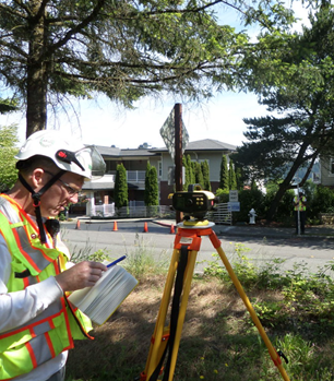 A person in a safety vest and helmet with field equipment, and a pen and paper capturing details about the road.