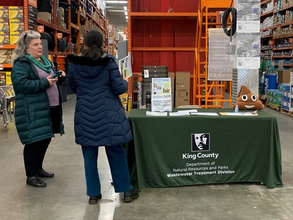 A community member with their back to the camera speaks with staff at Home Depot. A table to the right has informational materials on it.