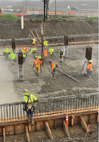 Construction staff in safety equipment laying concreate on the top of a stormwater vault surrounded by rebar and wood casings on a rainy day.