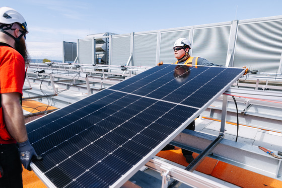 Two workers in helmets and orange vests position a large solar panel on a rooftop under a clear blue sky.