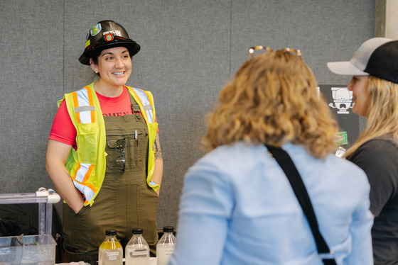 A female operator worker in a hard hat and safety vest smiling and speaking to group at an informational booth with sample bottles on the table.