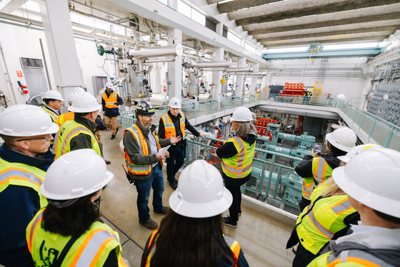 A large group in safety gear tours a massive industrial room with pipes, pumps, and engines. 
