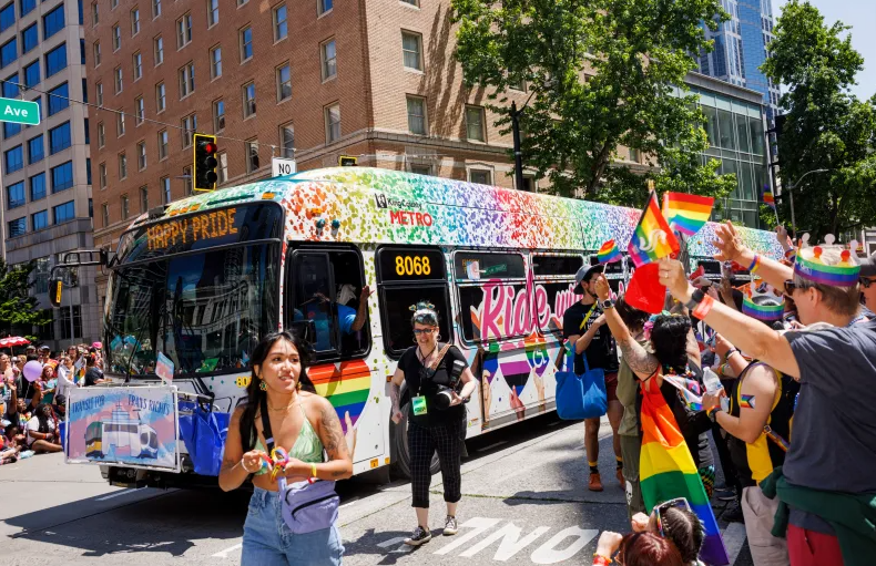 photo of Pride wrapped bus in past Pride parade with happy celebrating people all around 