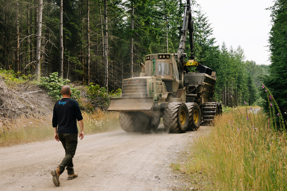  A man in a "Loop" shirt walks behind a large machine on a forest dirt road, surrounded by tall trees and wildflowers, creating a dusty scene.