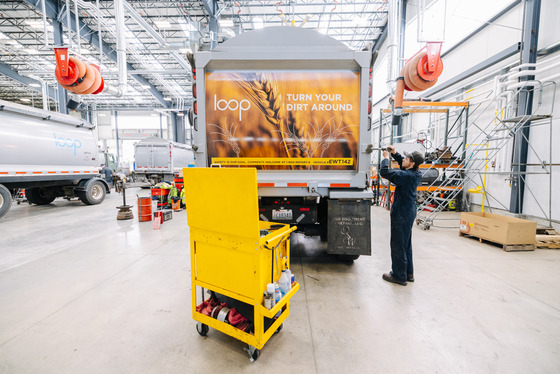 A worker in a warehouse checks on a truck with "Loop" branding and the slogan "Turn Your Dirt Around" on its back, surrounded by tools and trucks.