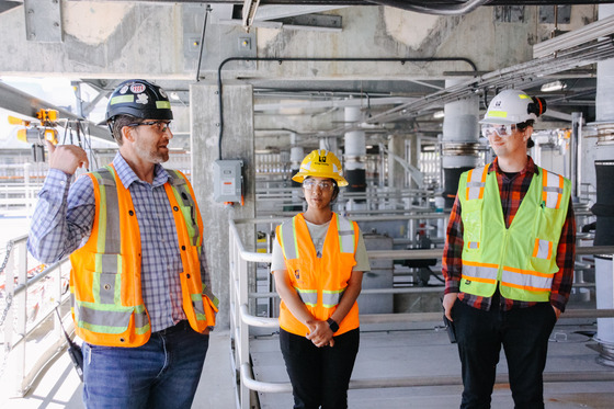 Three workers in safety gear and high-visibility vests converse in an industrial facility with pipes and equipment.