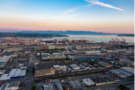 Aerial view of an industrial port with cranes and warehouses, set against mountains and a sunset sky.