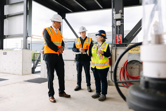  Three people in safety vests and hard hats converse at an industrial site.