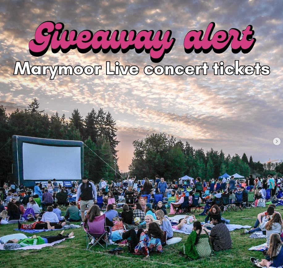 A crowd of people sit in the grass at an outdoor movie night in a park. 