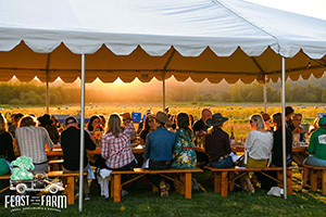 People sitting and eating at a long table under a canopy tent out in a agricultural field