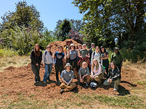 Group photo of a dozen joyful young adults in an open space surrounded with mature trees