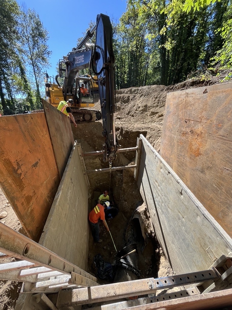 Crew members in personal protective equipment stands in the bottom of the trench between segments of excavated pipe.  