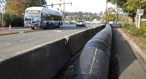 A black pipe on the ground runs parallel to a roadway and is protected by concrete barriers.