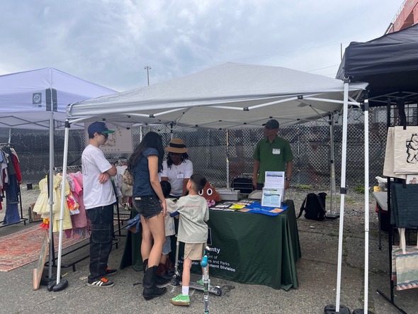 两位成年人和两名儿童站在户外帐篷下的信息展台前，一位 King County 工作人员正在分发项目资料和纪念品，另一位工作人员在旁边观察。