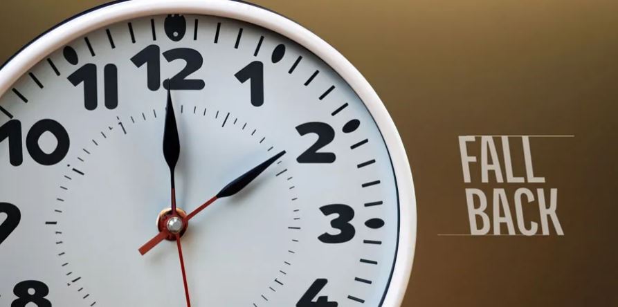 Image shows a clock face on a brown background, with text that reads "fall back"