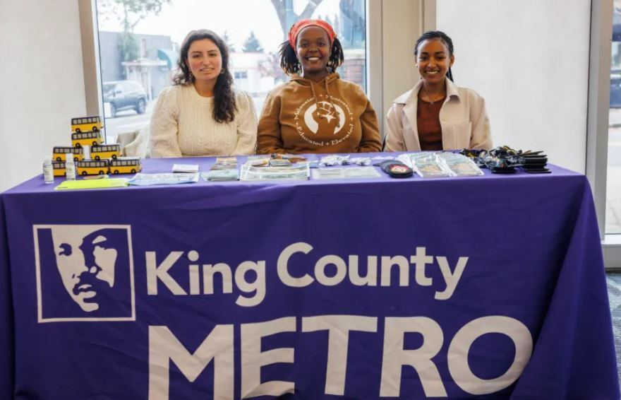 Three of Metro's Community Liasons are pictured smiling behind a table of transit swag