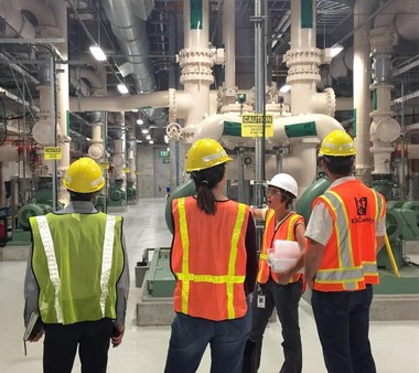 Four people on a tour wear personal protective equipment inside the plant. They are surrounded by pipes that connect to the ceiling.