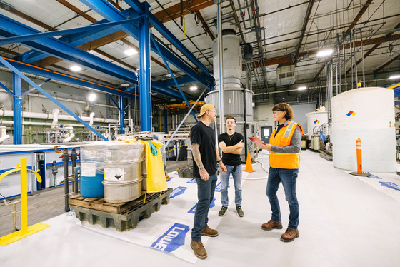 Three people stand in a factory near metal barrels, wearing safety gear and casual work attire, surrounded by industrial equipment and supplies.