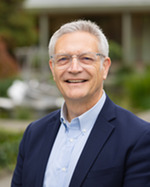 A person with short gray hair, wearing glasses, a light blue shirt, and a navy blazer, smiles in an outdoor setting with greenery in the background.