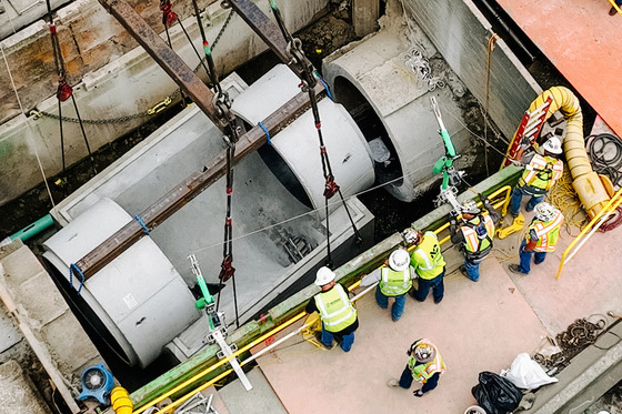  Construction crew installs large concrete pipes using machinery. Workers in safety gear stand near in an industrial setting.