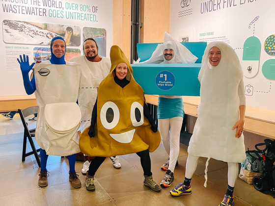 Five people in costumes of wastewater-related items, like a toilet, poop emoji, and wipes, pose indoors.