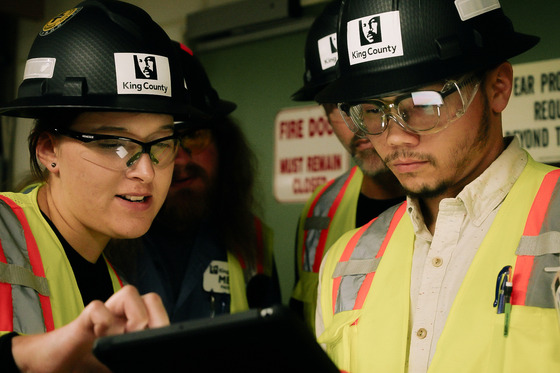 Two people wearing King County hard hats, safety glasses, and high-visibility vests review a tablet while others stand nearby in a work setting