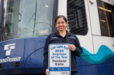 Image of Pardeep Kaile smiling in from of light rail holding sign / award