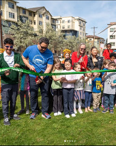 A group of people cut a green ribbon while standing in the grass. Trees and apartments are behind them