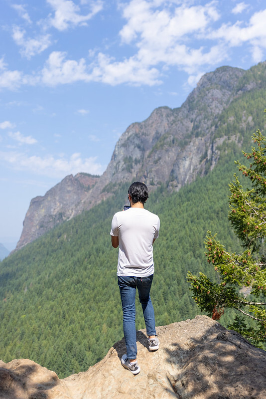 A person stands with their back to the camera while looking out a nearby mountain covered in trees and rocky outcrops