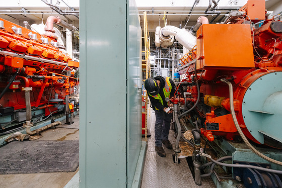 Worker in safety gear inspects an engine inside a wastewater treatment plant, surrounded by piping and control equipment.