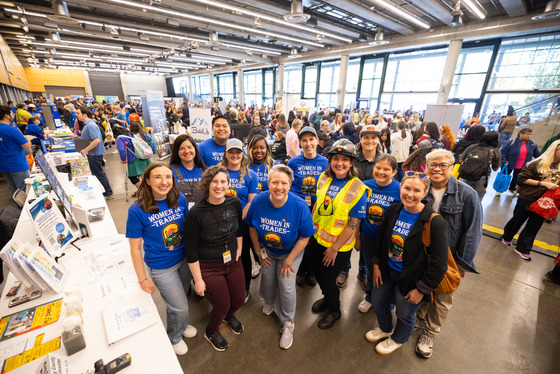 Group of King County staff wearing “Women in Trades” shirts pose at a busy career fair booth, surrounded by attendees and info displays.