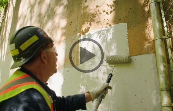 Worker wearing safety gear paints a concrete wall with a roller, covering old, weathered surface with a fresh coat.