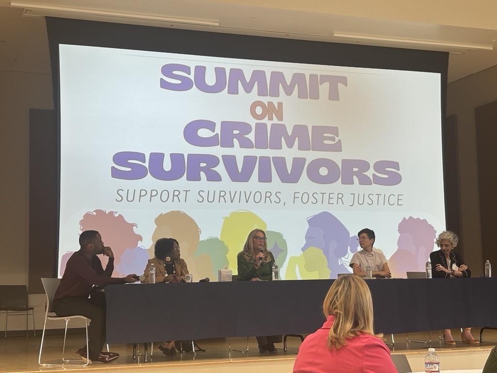A panel of speakers sit in front of a projected image saying "Summit on Crime Survivors, Support Survivors, Foster Justice"
