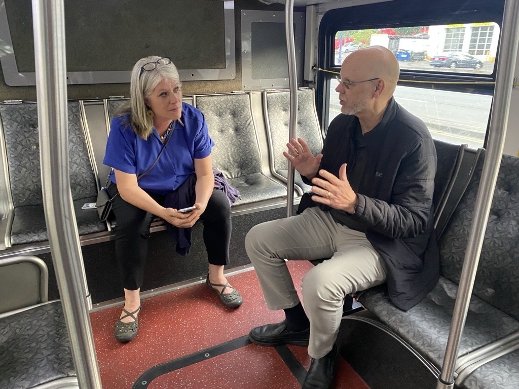 Councilmember Balducci in a blue shirt sits on a Metro bus listening to a man in a black jacket speak
