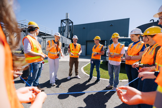 A group of people in hard hats and safety vests stands in a circle outside, holding a rope and listening to a speaker.