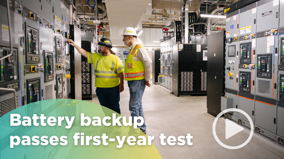 Workers in safety gear inspect electrical control panels inside a facility, with the text "Battery backup passes first-year test"