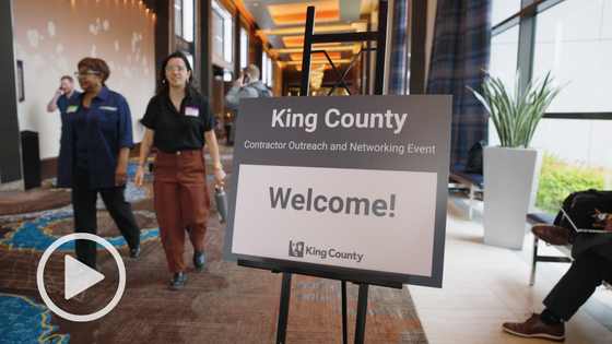 ChatGPT said: A sign reads "King County Contractor Outreach and Networking Event Welcome!" as people walk through a hallway at the venue.