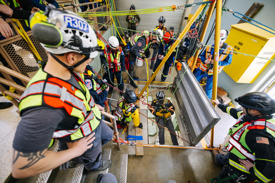Firefighters in safety gear and helmets practice a confined space rope rescue drill inside a stairwell, using ropes and harnesses.