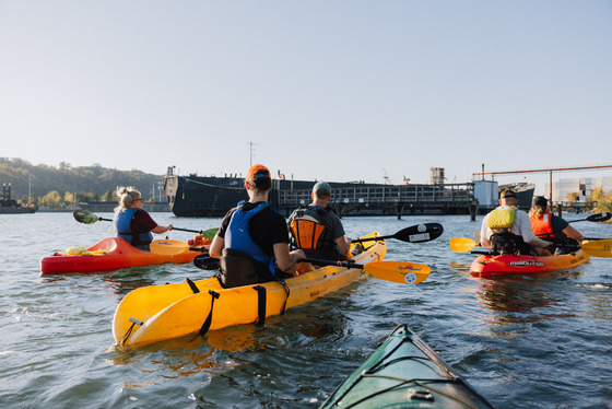A group of kayakers paddle along the Duwamish River near an industrial pier.
