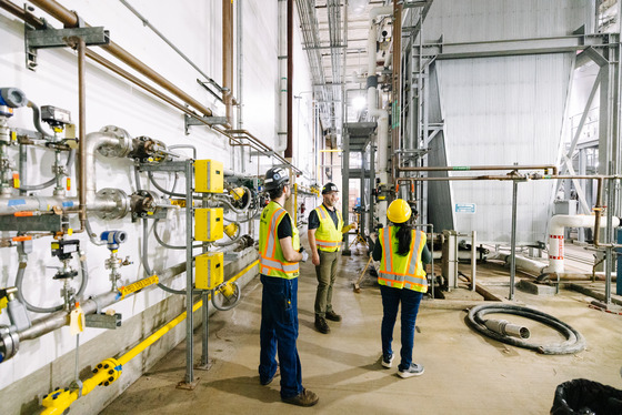 Three workers in safety vests and helmets stand inside a treatment plant, surrounded by pipes and equipment.