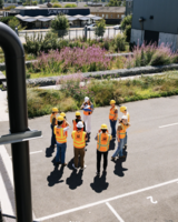 A group of people in hard hats and safety vests gather outdoors for a tour near landscaping and buildings.